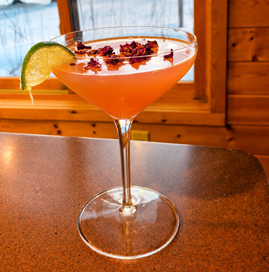 Pink cocktail in a martini glass with a lime wedge and rose petals